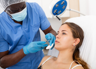 Professional male beautician performing face contouring injections to female patient at aesthetic...