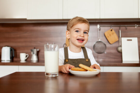 little boy of 2 years old sits in the kitchen at the table and eats cookies with milk and smiles