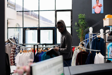 African american robber trying to steal clothes from shopping mall, looking at hangers full with...