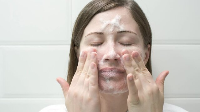 Young woman applying cleansing foam to her face close-up on white tiles background.