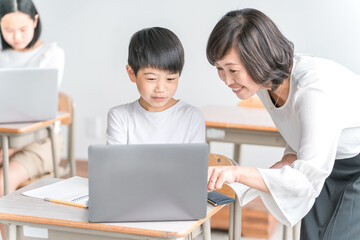 学校の教室でノートパソコンを使いながら勉強する男子小学生と女性講師（教育・先生・授業・家庭教師）
