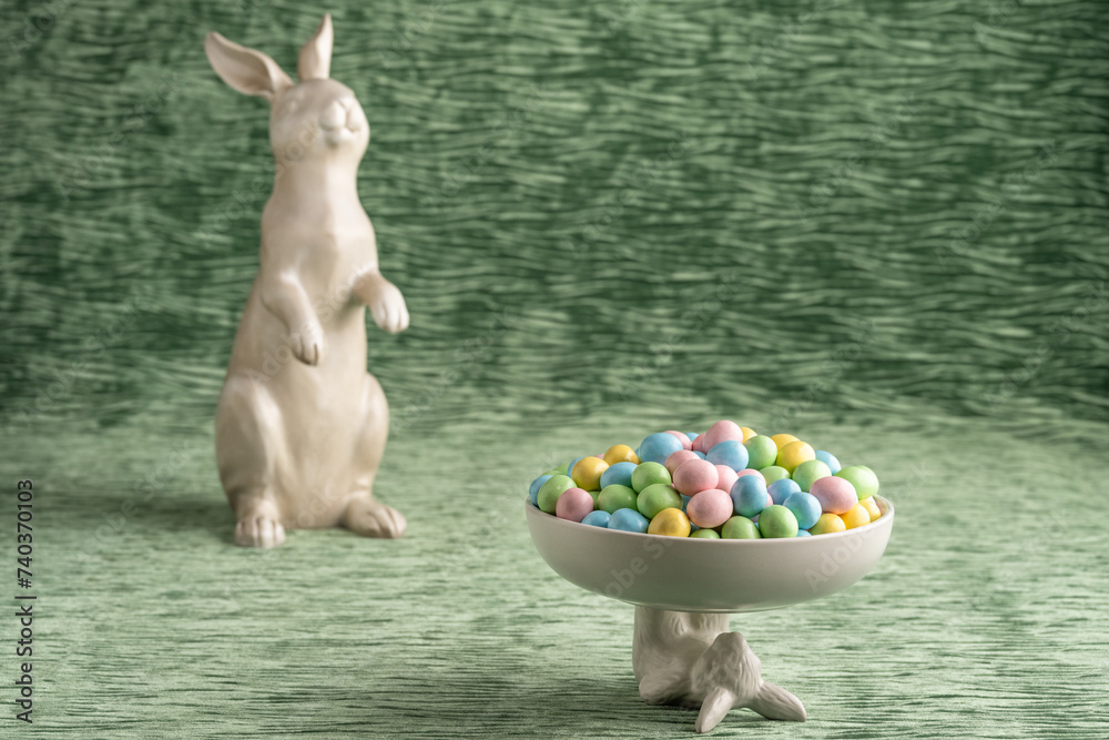 Wall mural Decorative white bowl held up by the Easter Bunny full of candy coated pastel easter eggs, with large white Easter Bunny in background, on a textured green velvet background, Happy Easter
