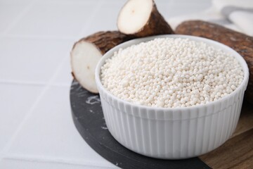 Fototapeta na wymiar Tapioca pearls in bowl and cassava roots on white tiled table, closeup. Space for text