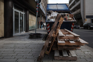 Wooden pallets stacked haphazardly in the foreground. Behind, a large dumpster overflows with...