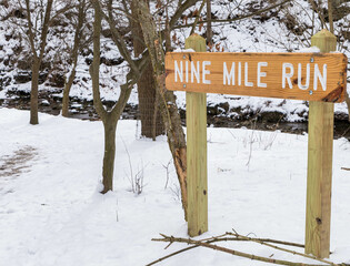 A wooden sign engraved with Nine Mile Run, a stream in Frick Park, an urban park in Pittsburgh, Pennsylvania, USA as seen on a sunny winter day
