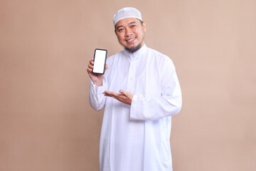 Asian muslim man in white robe showing phone screen over beige background