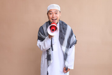 Young Asian Man Wearing Muslim Robe Announcing News Using Megaphone Isolated on Beige Background