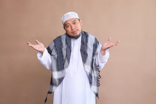 Confused Asian muslim man shrugging shoulders on beige background