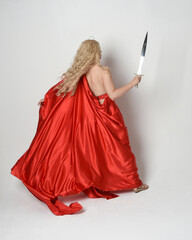 Full length portrait of beautiful blonde model dressed as ancient mythological fantasy goddess in flowing red silk toga gown, crown.  back view, walking away from camera  holding a sword weapon - obrazy, fototapety, plakaty