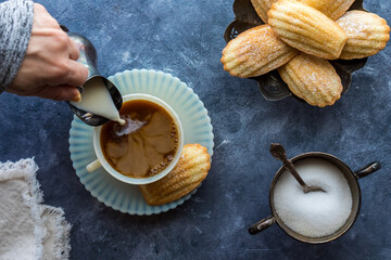 Cream being poured into a cup of coffee served with French Madeleines.