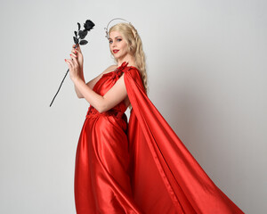 Close up portrait of beautiful blonde model wearing flowing red silk toga gown and crown, dressed as ancient mythological fantasy goddess. holding a rose flower isolated  studio background.
