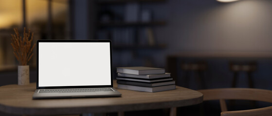 A white-screen laptop computer mockup and books on a table in a modern dark room.