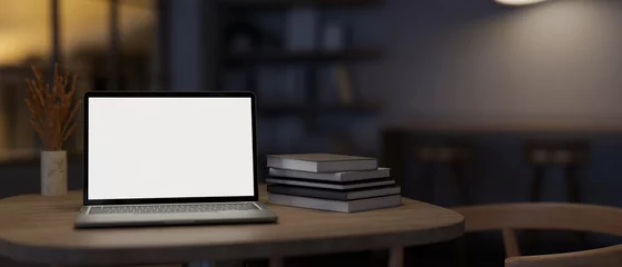  A white-screen laptop computer mockup and books on a table in a modern dark room. © bongkarn