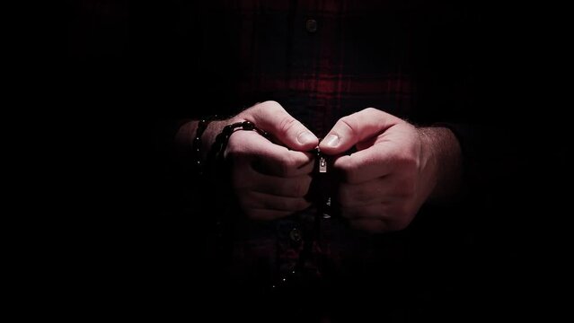 A man's hands wrapped with a wooden Rosary comes out of the dark into the light with praying hands to Jesus.