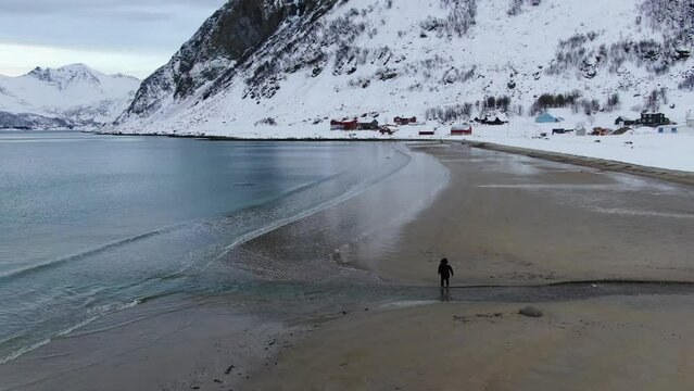 Drone aerial view in Norway flying over a person running in circles on a beach surrounded by white snow mountains in Tromso