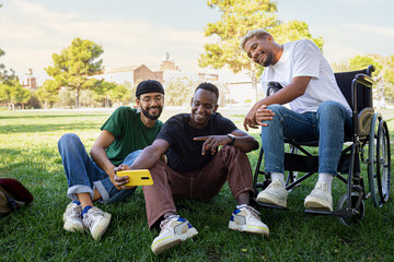 Young Black man in a wheelchair and his male friends taking selfie with phone sitting on the grass...