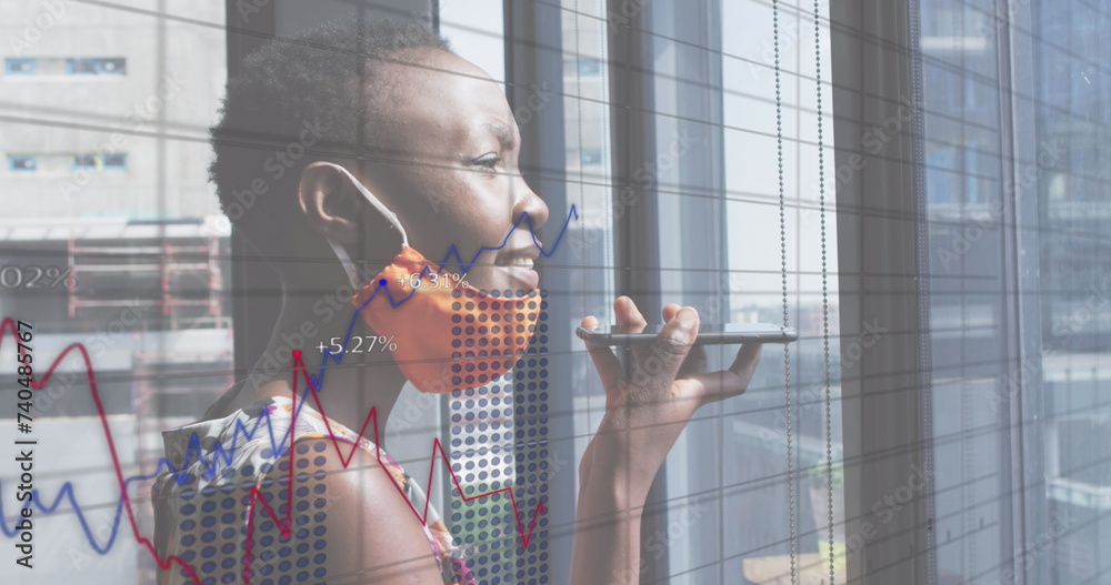 Wall mural Image of financial data processing over businesswoman with face mask using smartphone in office