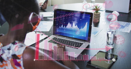 Image of financial data processing over businesswoman with face mask using laptop in office