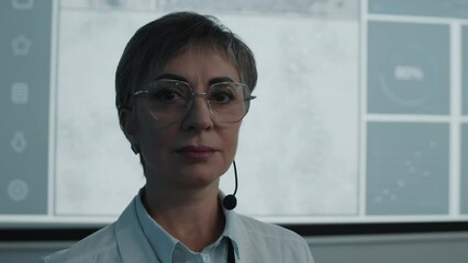 Portrait of female speaker with microphone headset wearing white coat and eyeglasses and posing for camera in front of projection screen during medical conference