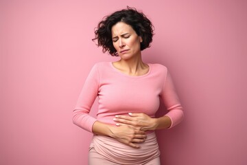 
Photography a middle-aged woman holding her belly with a worried expression, against a solid pastel pink background, portraying the discomfort of her stomach pain - obrazy, fototapety, plakaty