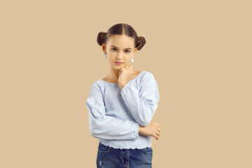 Adorable kid in casual summer outfit. Child posing on beige fashion studio background. Pretty Caucasian girl with hair buns standing with her hand on chin and looking at camera with pensive expression