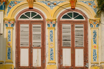 The warm afternoon sun bathes a centuries-old shophouse, revealing the timeless elegance of two vintage wooden louvred windows, adorned with intricate floral carving in Malacca heritage site.