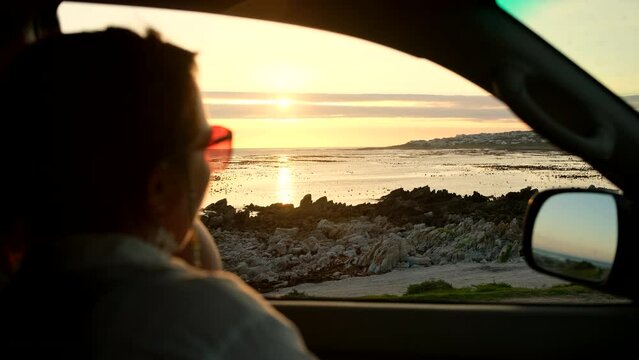Woman in car points to something in sea while sun is setting over ocean