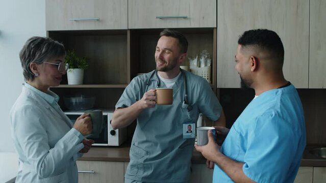 Company of hospital workers having discussion and drinking from mugs while gathering for tea break in staff room