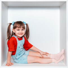 A little girl sits in a box, on a white background. The girl is 3 years old. The child climbed into the white cabinet. She smiles beautifully. Happy childhood.
