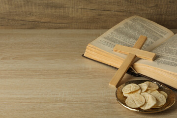 Bread on golden plate and wooden cross on book on wooden background, space for text
