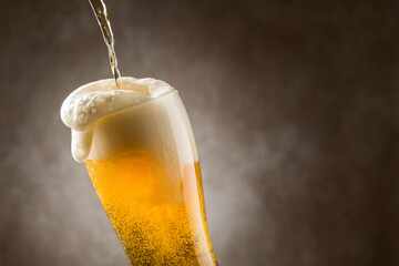 Pouring lager beer into glass with foam out on dark background.