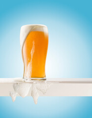 Glass of blonde beer with foam out on white shelf on blue background.