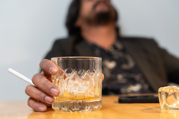 Close-up of a hand holding a glass of liquor. A disheveled man sits in his living room, surrounded...