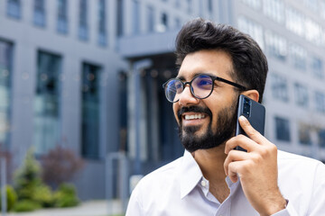 Cheerful young professional engaging in a phone conversation with a modern smartphone against the backdrop of urban architecture.