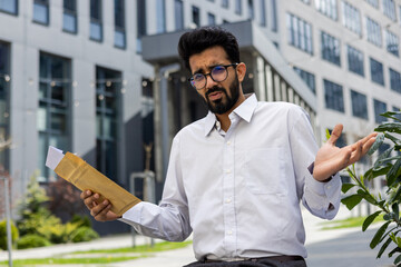 Perplexed young male professional holding a brown envelope with a bewildered expression in an urban setting.