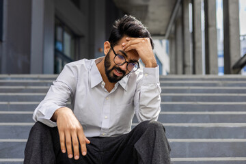 A stressed businessman in despair sits on outdoor steps, expressing worry and frustration,...