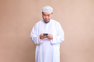 Religious Asian man in white robe and skullcap using mobile phone over beige background