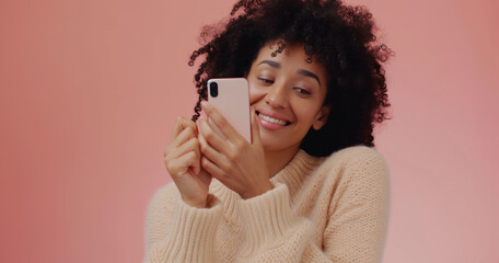 Beautiful young black woman texting on cell phone and giggling at flirty text, pink background - obrazy, fototapety, plakaty