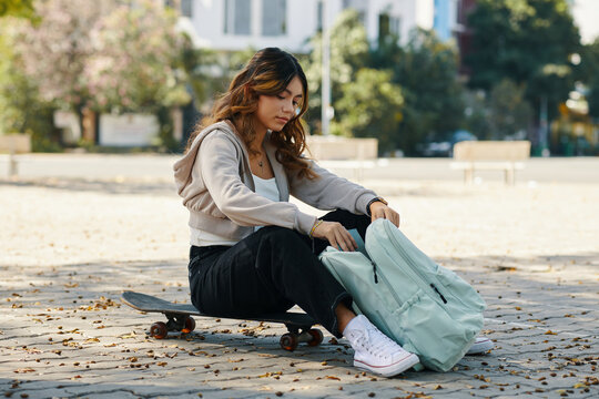 Teenage girl sitting on skateboard and taking book out of backpack