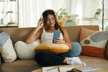 Smiling girl sitting on couch and watching music videos on smartphone