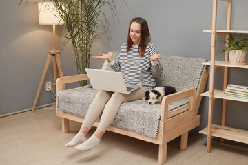 Uncertain Caucasian woman wearing striped shirt sitting on sofa with her puppy dog having video...