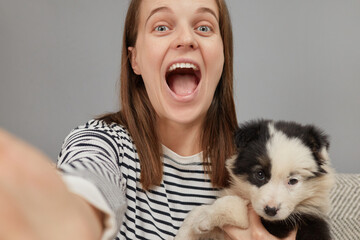 Beautiful young adult cheerful woman taking selfie on smartphone with her dog exclaiming with excitement and happiness keeps mouth widely opened making point of view photo for her network page