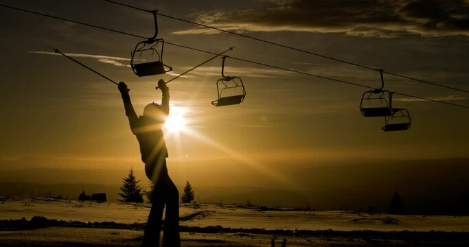 Silhouette of a female skier against sunset sky beneath chairlift. Girl rasing arms with ski poles. 120 fps on cinema camera