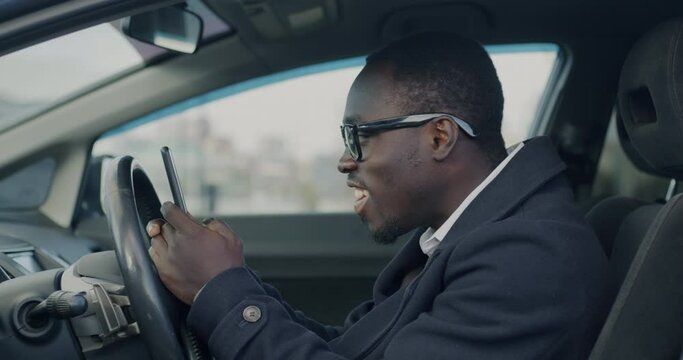 African American entrepreneur using smartphone texting and driving car in urban street in modern city. Transportation and mobile communication concept.