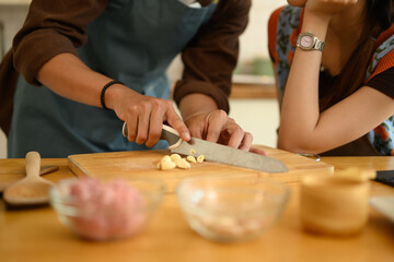 Man in apron chopping garlic on wooden board while preparing meal with his wife in the kitchen