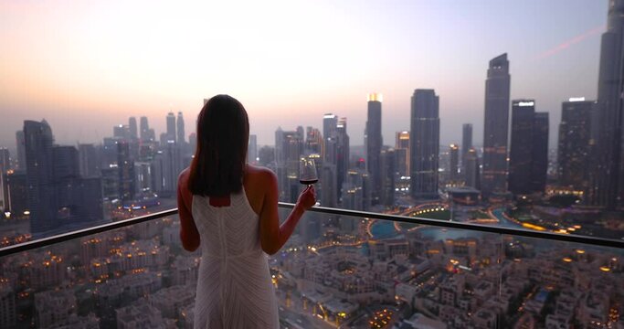 A beautiful luxury woman in a white dress enjoys the sunset view behind the modern skyline of Downtown Dubai, UAE, with a drink