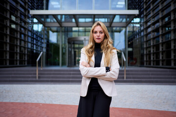 Fototapeta premium Stylish adult business beautiful blonde woman in suit standing in corporate office area. Empowered Caucasian female posing confident looking serious at camera outdoor. Copy space building background