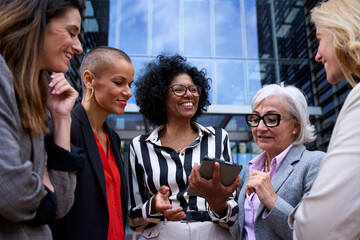 Team diverse ages and race business women conversing cheerful standing outside office building. Professionals successful positive mature females talking about commerce looking and using tablet in hand - obrazy, fototapety, plakaty