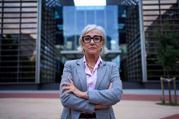 Successful and serious middle-aged business gray woman with arms crossed at workplace looking at...