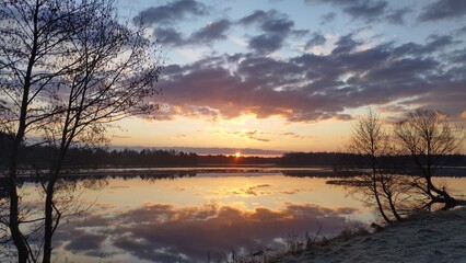 The sun rises from behind the forest on the far side of the lake and colors the sky with bright...